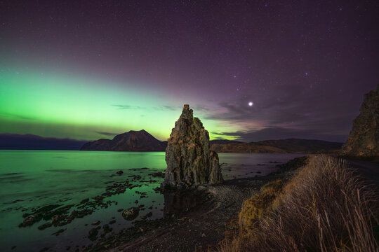 View of the iconic rock formation rising from the shoreline under a vibrant display of the aurora borealis illuminating the night sky in Unalaska, Alaska, United States.