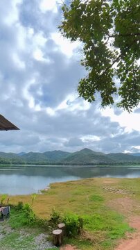 Vertical timelapse of clouds moving over a mountain lake surrounded by forested hills, calm water and a grassy shoreline, capturing a scenic natural landscape.