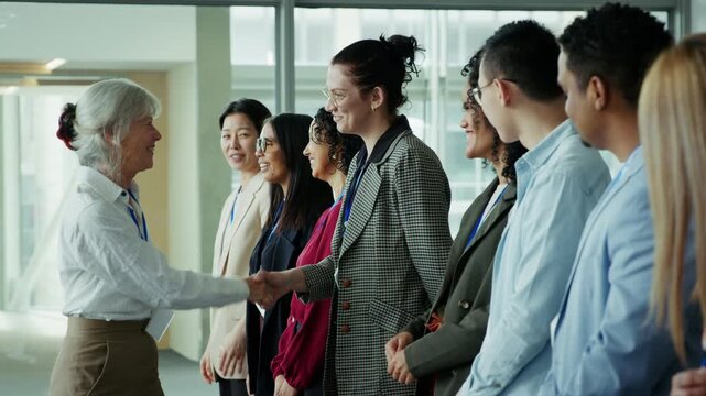A senior business professional shakes hands and greets a line of smiling, enthusiastic new colleagues at a professional conference or corporate event, fostering connections and building team rapport.