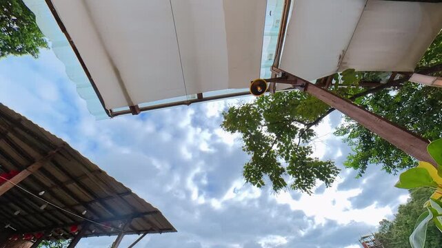 Timelapse of clouds moving across a blue sky framed by tropical tree branches, rooftops and a covered outdoor structure, capturing changing weather and natural scenery.
