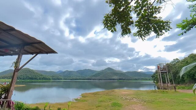 Timelapse of clouds moving above a mountain lake surrounded by forested hills, with a grassy shoreline and dramatic sky creating a scenic natural landscape.