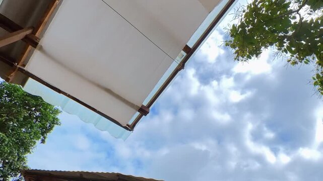 Timelapse of clouds moving across a blue sky, framed by tropical foliage and rooftop structures, capturing changing weather and natural outdoor scenery.