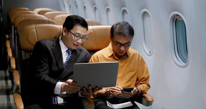 Asian businessman and senior male passenger review laptop and smartphone beside airplane window inside modern aircraft cabin, representing business travel, mobile work and onboard productivity.