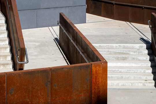 Weathered steel walls frame concrete steps in modern urban architecture where rusted surfaces railings and geometry define a sculptural stairway