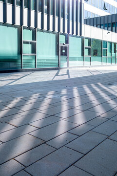 Modern glass building casts long geometric shadows across an empty urban pavement where facade lines create a graphic architectural pattern