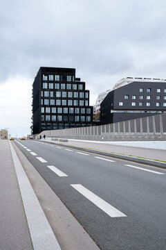 Empty urban road leads toward modern buildings beneath an overcast sky where lane markings barriers and perspective shape a quiet transportation scene