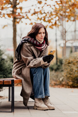 Obraz premium Young woman in coat and scarf sitting on a bench in autumn checking her phone with a worried expression