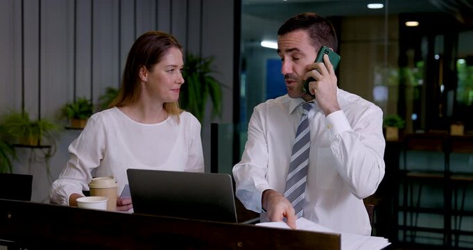 Caucasian business colleagues coordinate work in modern coworking office as businessman talks on smartphone, then checks mobile screen with businesswoman before both continue laptop collaboration.