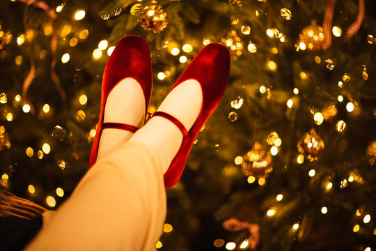 Red ballet flats worn by a person with light-colored pants, positioned in front of a decorated Christmas tree with golden ornaments and soft glowing lights