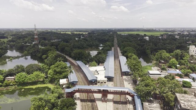 Forward-moving drone footage showcases Poradah Junction railway station in Kushtia, Bangladesh, with multiple tracks, covered platforms, a pedestrian footbridge, and lush green surroundings.