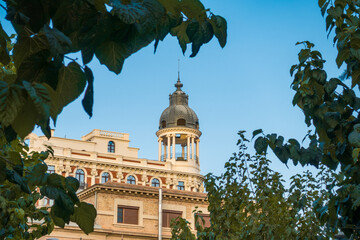 Classic building tower dome framed by green tree leaves against clear blue sky © Mariya