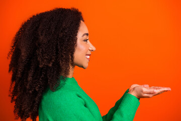 Young woman in green sweater smiling in profile with palm up presenting copy space on vibrant orange background, cheerful portrait and offering gesture © deagreez
