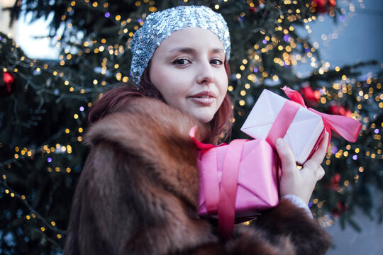Young woman with red hair wearing a silver headband holds pink and white wrapped gifts in front of a decorated Christmas tree with lights and ornaments