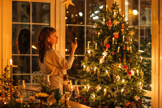 Woman taking photo of decorated Christmas tree with lights and ornaments in cozy indoor setting, candles and festive decorations visible in the background