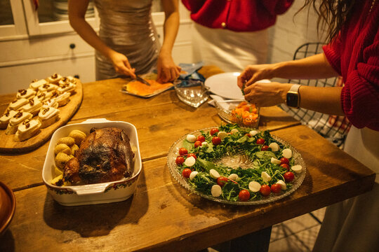 Group of people preparing festive meal with roasted meat, vegetable platter, and appetizers on wooden table in cozy kitchen setting during holiday gathering