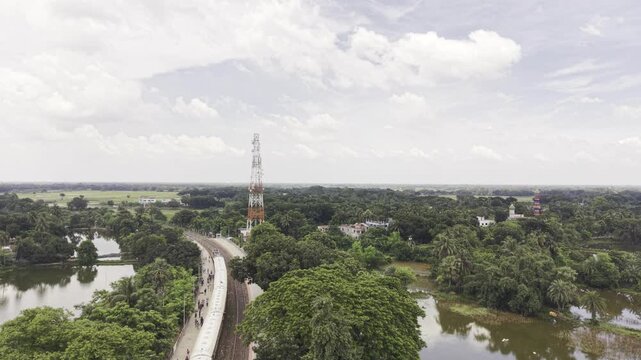Drone footage captures a passenger train leaving Poradah Junction railway station in Kushtia, with railway platforms and tracks extending through a green countryside surrounded by ponds and trees.