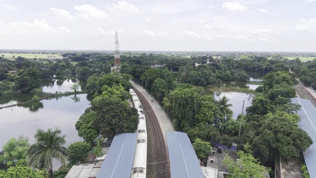 Aerial drone footage moves forward over Poradah Junction in Kushtia, Bangladesh, revealing multiple railway tracks, covered platforms, a pedestrian footbridge, and dense greenery around the station.