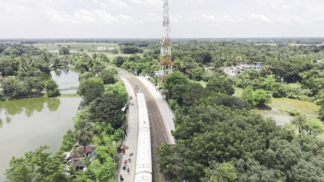 Aerial drone footage shows a passenger train departing Poradah Junction in Kushtia, Bangladesh, moving between railway platforms through a lush rural landscape with ponds, trees, homes, and farmland.