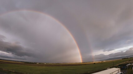 Obraz premium Rainbow arcs across a stormy sky over a wide green field.