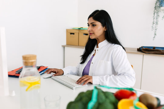 Mid adult Indian female dietitian using computer at desk. Nutrition professional working beside healthy food for consultation. Nutrition and wellness concept.