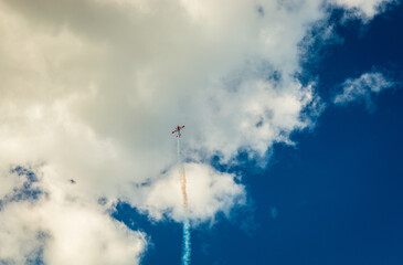 Small aerobatic stunt plane climbing vertically into cloudy blue sky with white smoke trail during airshow © Damian