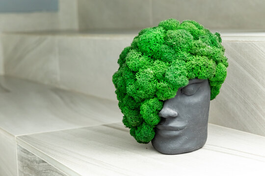 Decorative black sculpture shaped like a human head features lush green moss that mimics hair, placed on a smooth light-colored stone surface, creating a modern artistic display