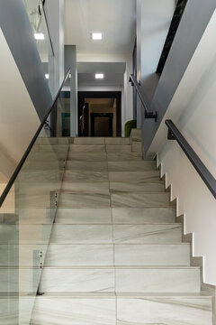 Modern interior staircase featuring polished stone steps and a glass railing, leading to a well-lit hallway with minimalistic design elements in a contemporary building