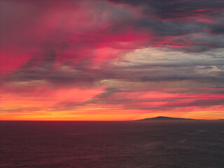 Obraz premium Intense crimson and orange sunset clouds spread across the sky above the Pacific Ocean off the Southern California coast, creating a dramatic fiery atmosphere over the distant shoreline and horizon.