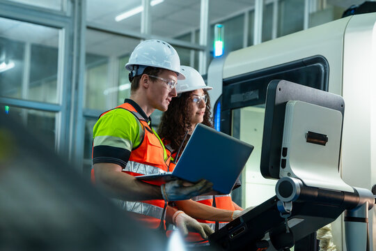 Team of male and female industrial engineers in safety gear using a laptop to program and control a CNC machine on a modern manufacturing factory floor. Teamwork and smart industry concept.