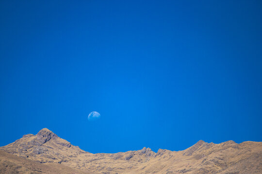 View of the Sacred Valley mountain range under a clear blue sky with a visible moon in Urubamba Province, Cusco, Peru.