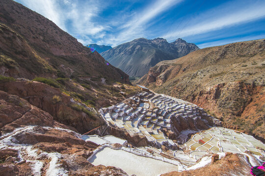 View of the Salt Mine of Maras with its terraced salt evaporation ponds nestled in the rugged Andean mountains under a bright blue sky in Urubamba, Cusco, Peru.