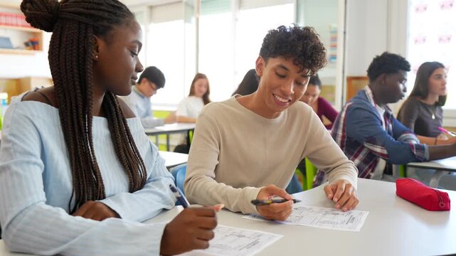 Two multiracial high school students sitting at their desk, discussing and working together on a school test, helping each other with the answers in a brightly lit modern classroom