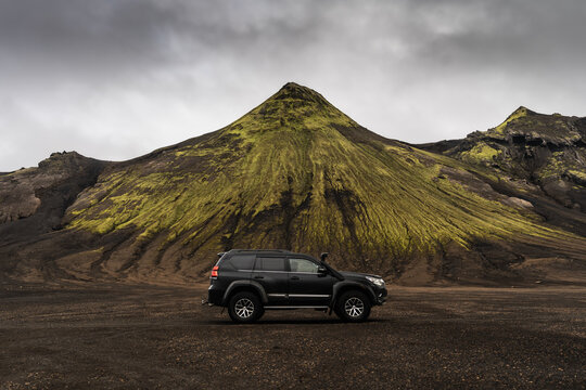 View of a black SUV parked on dark volcanic soil in front of a prominent moss-covered mountain in the Icelandic Highlands, Skaftarhreppur, Iceland.