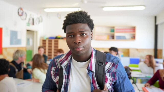 Young african american male student with a backpack looking directly at the camera while standing in a high school classroom with other multiracial classmates sitting at their desks