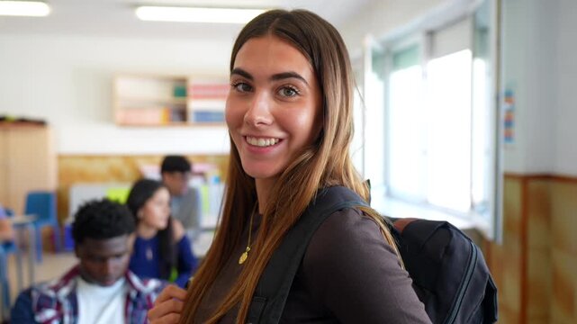 Portrait of a beautiful young female student with a backpack looking at the camera and smiling, with her multi ethnic classmates studying in the background inside a high school classroom