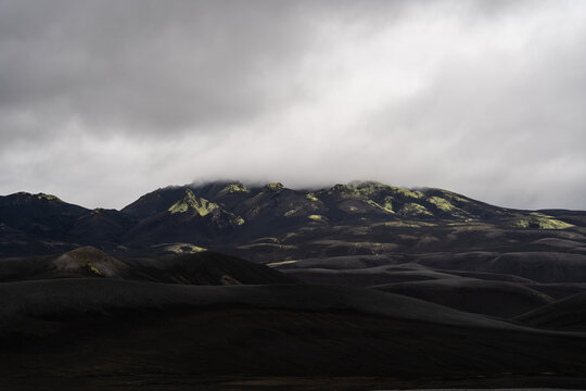View of the Icelandic Highlands featuring dark volcanic terrain with patches of vibrant green moss under a heavy overcast sky in Skaftarhreppur, Iceland.
