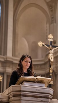Woman lector reading gospel from ambo in church during catholic mass. Female reader proclaiming word of god at pulpit with crucifix in background.