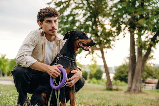 Young man crouches beside a Doberman dog on a grassy field in a park, holding a purple dog leash with trees and a cloudy sky in the background
