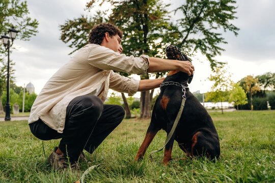 Young man crouching on grass petting a Rottweiler dog in a park with trees and cloudy sky in the background, showcasing a moment of companionship