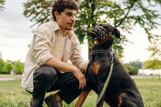Young man kneeling beside a Rottweiler dog wearing a muzzle in a green park with trees and a cloudy sky visible in the background