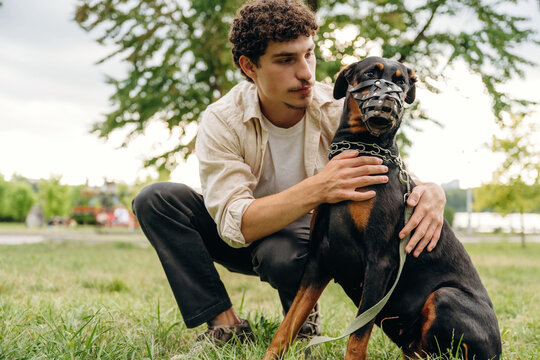 Young man with curly hair kneeling beside a Doberman dog wearing a muzzle in a green park setting with trees and grass in the background