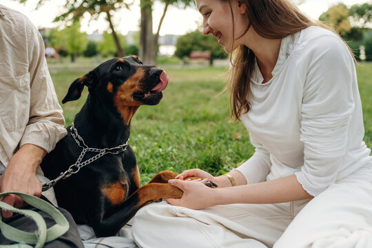 Woman in white shirt interacts with a Doberman dog on a blanket in a park, with green grass and trees in the background creating a serene outdoor setting