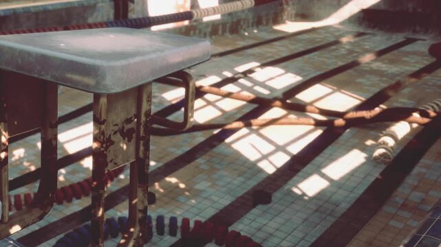 A diving board stands at the edge of an unused swimming pool. Sunlight pours in, creating patterns on the tiled floor. Lane ropes are visible beside the pool area.