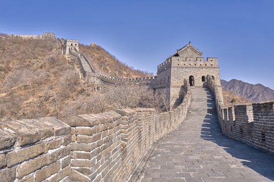 View of the Great Wall of China with its stone path, ancient watchtowers, and rugged hills under a clear blue sky in Beijing, China.