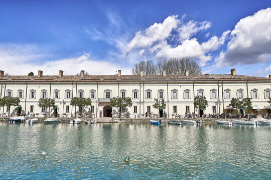 View of Lake Garda with a historic building along the waterfront, small boats moored in the clear water, and trees under a bright blue sky in Veneto, Italy.