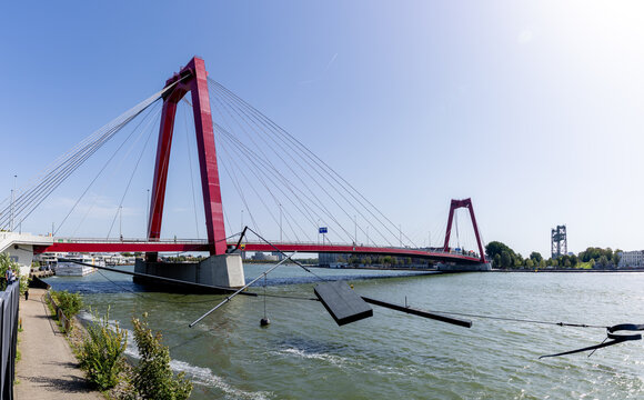 View of the Willemsbrug, a striking red cable-stayed bridge spanning the Nieuwe Maas river under a clear blue sky in Rotterdam, South Holland, Netherlands.