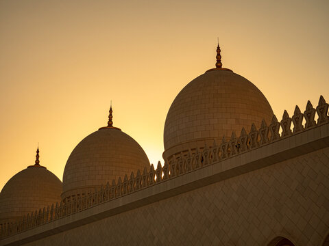 View of the white marble domes and intricate parapets of the Sheikh Zayed Grand Mosque silhouetted against a warm golden sunset sky in Abu Dhabi, Abu Dhabi, United Arab Emirates.