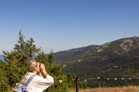 Woman looks through binoculars at Śnieżne Kotły mountain range from Szrenica peak viewpoint. Concept of scenic nature observation and mountain panorama viewing during solo hiking in Karkonosze.