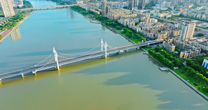 Aerial view of a white suspension bridge spanning a wide river alongside dense urban skyscrapers under bright sunlight.