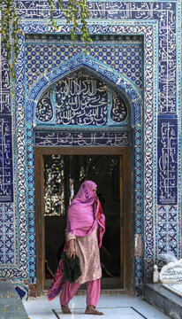 View of a woman in a pink headscarf walking past the ornate blue-tiled entrance of the Tomb of Shah Yusuf Gardezi in Multan, Punjab, Pakistan.
